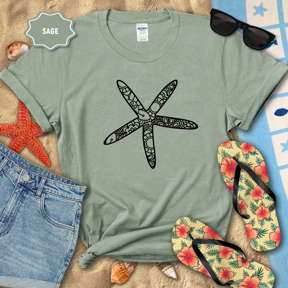 Sage green t-shirt with a starfish design on a sandy surface with beach accessories.