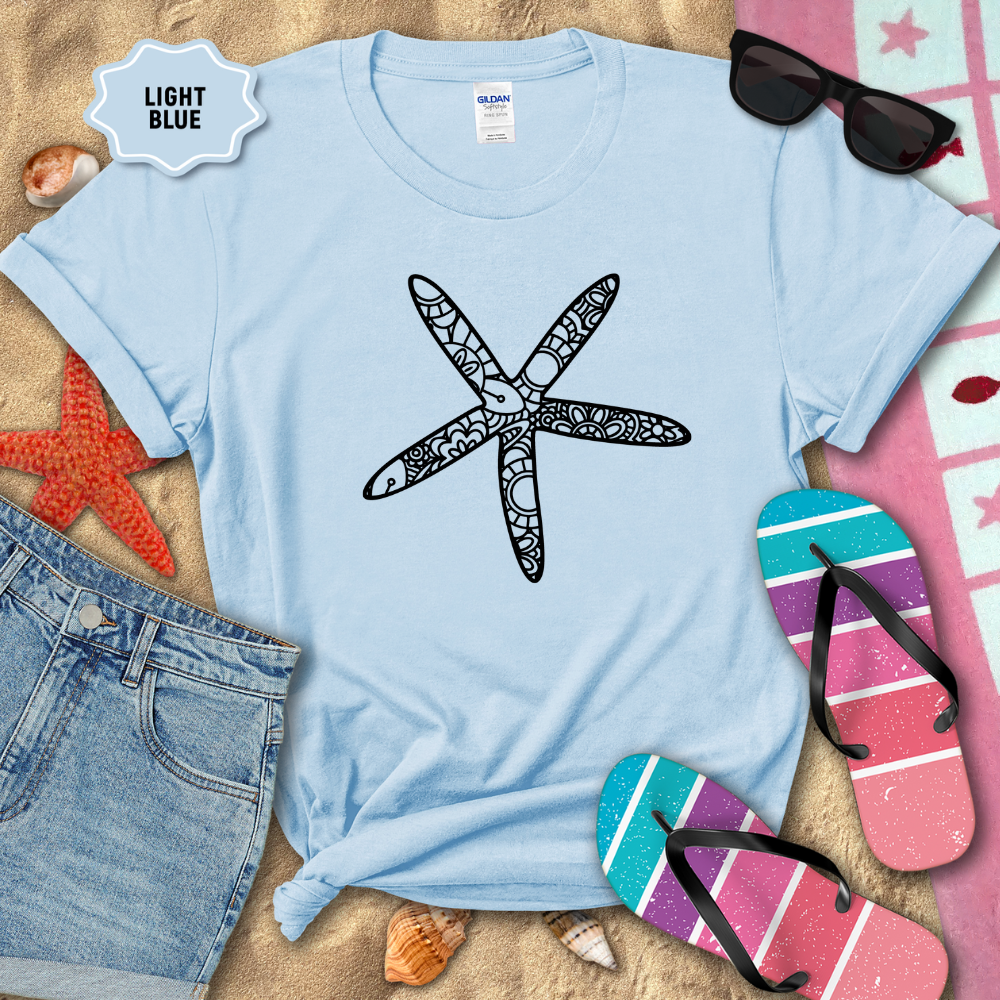 Light blue t-shirt with a starfish design on a sandy background with beach accessories.