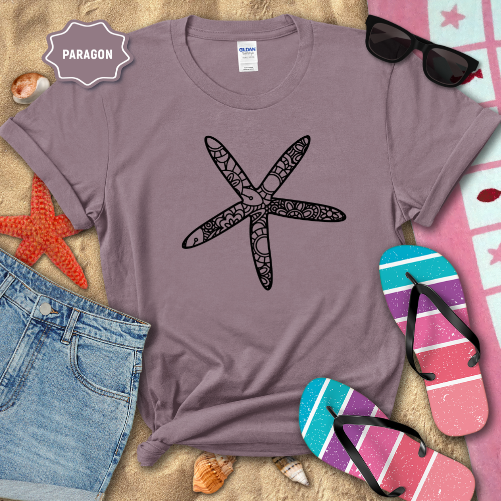 Purple t-shirt with a starfish design on a sandy background with beach accessories.