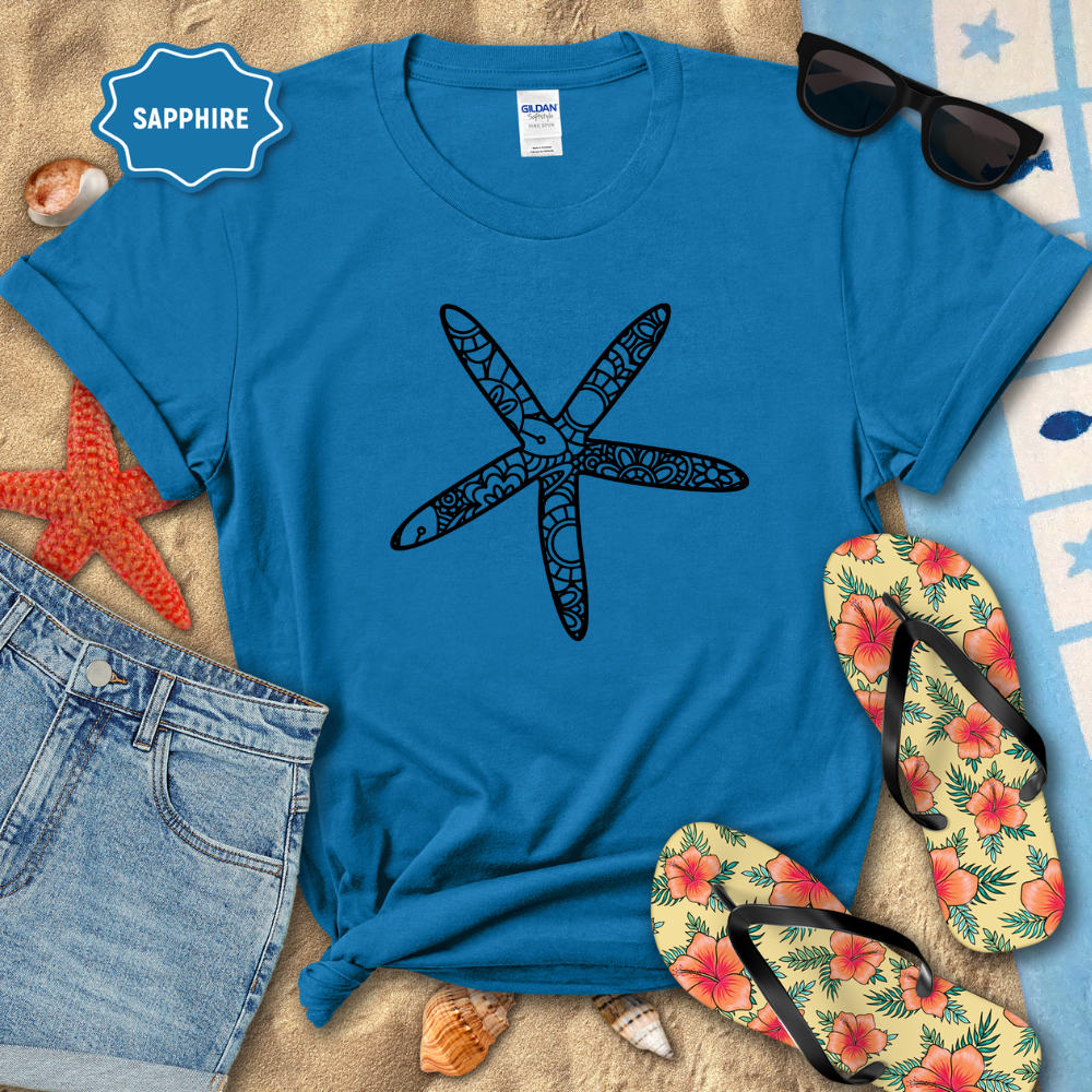 Blue t-shirt with a starfish design on a sandy surface with flip-flops and sunglasses.