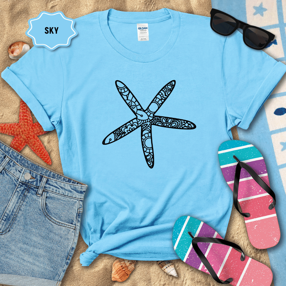 Light blue t-shirt with starfish design on a sandy surface with beach accessories.