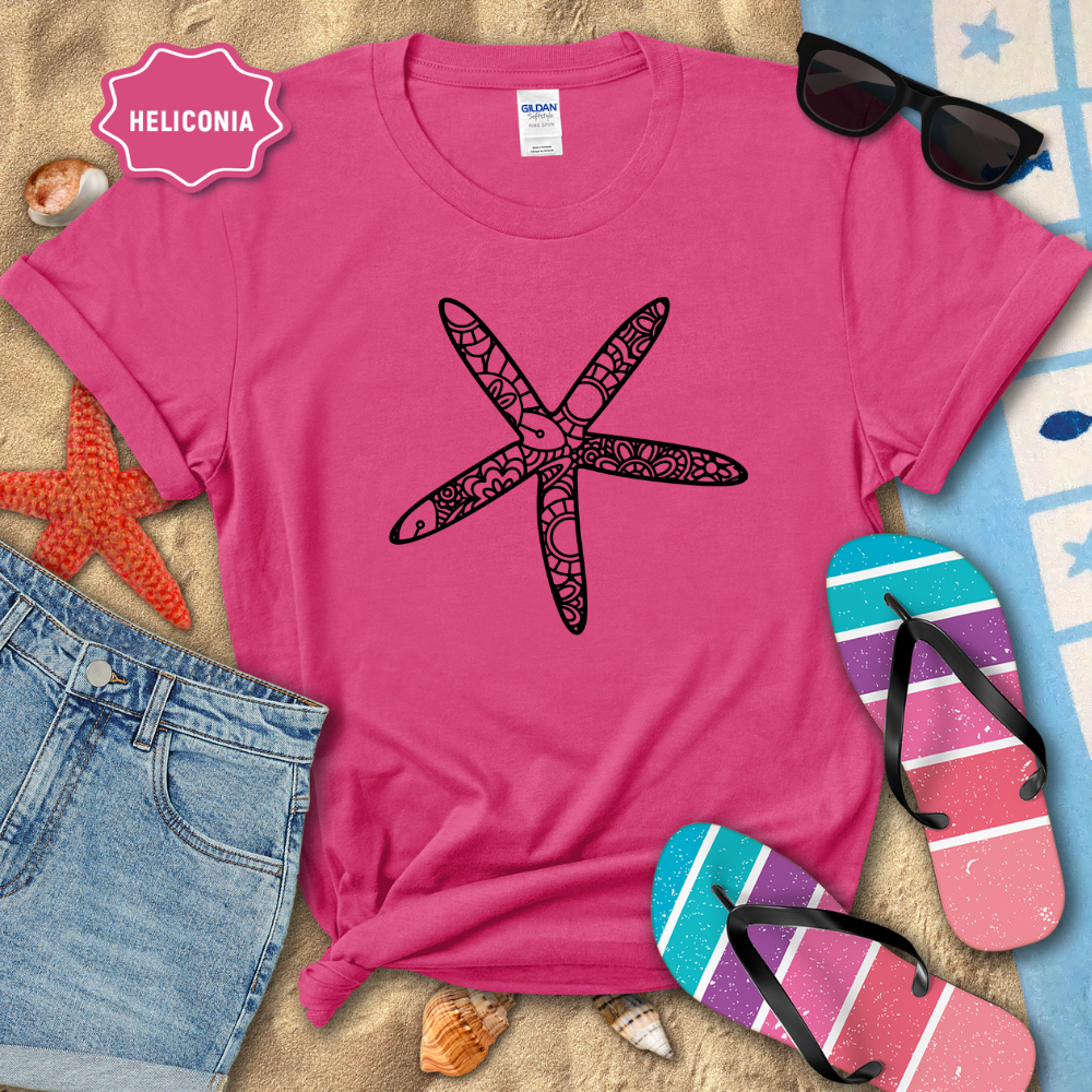 Pink t-shirt with a black starfish design on a sandy surface with beach accessories.