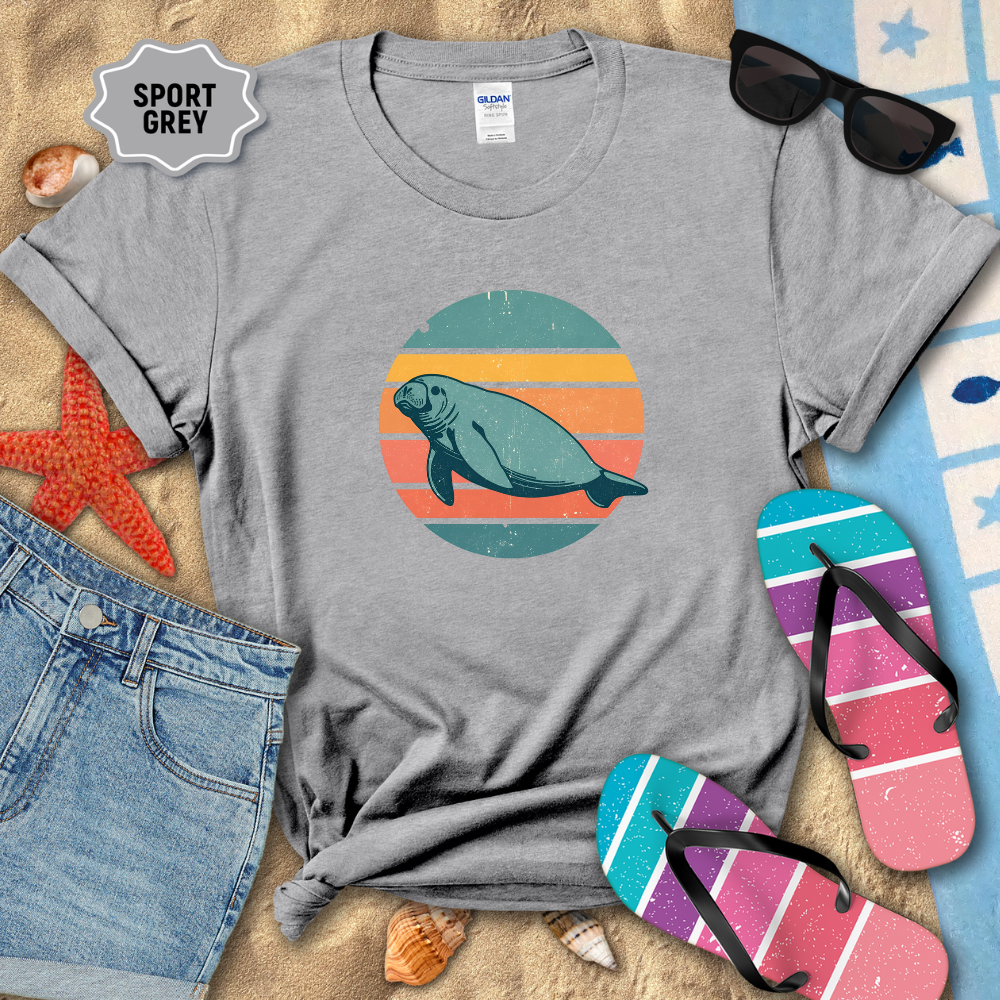 Retro sunset manatee graphic t-shirt in a sport gray color, styled on beach sand with seashells, flip-flops, sunglasses, and denim shorts. The shirt features a circular vintage sunset with teal, coral, and orange stripes and a teal manatee illustration in the center.