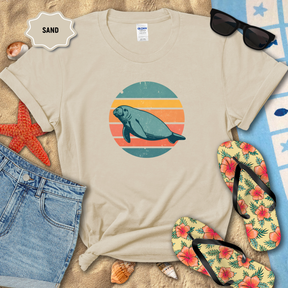 Retro sunset manatee graphic t-shirt in a natural color, styled on beach sand with seashells, flip-flops, sunglasses, and denim shorts. The shirt features a circular vintage sunset with teal, coral, and orange stripes and a teal manatee illustration in the center.