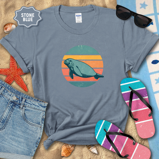 Retro sunset manatee graphic t-shirt in a stone blue, styled on beach sand with seashells, flip-flops, sunglasses, and denim shorts. The shirt features a circular vintage sunset with teal, coral, and orange stripes and a teal manatee illustration in the center.