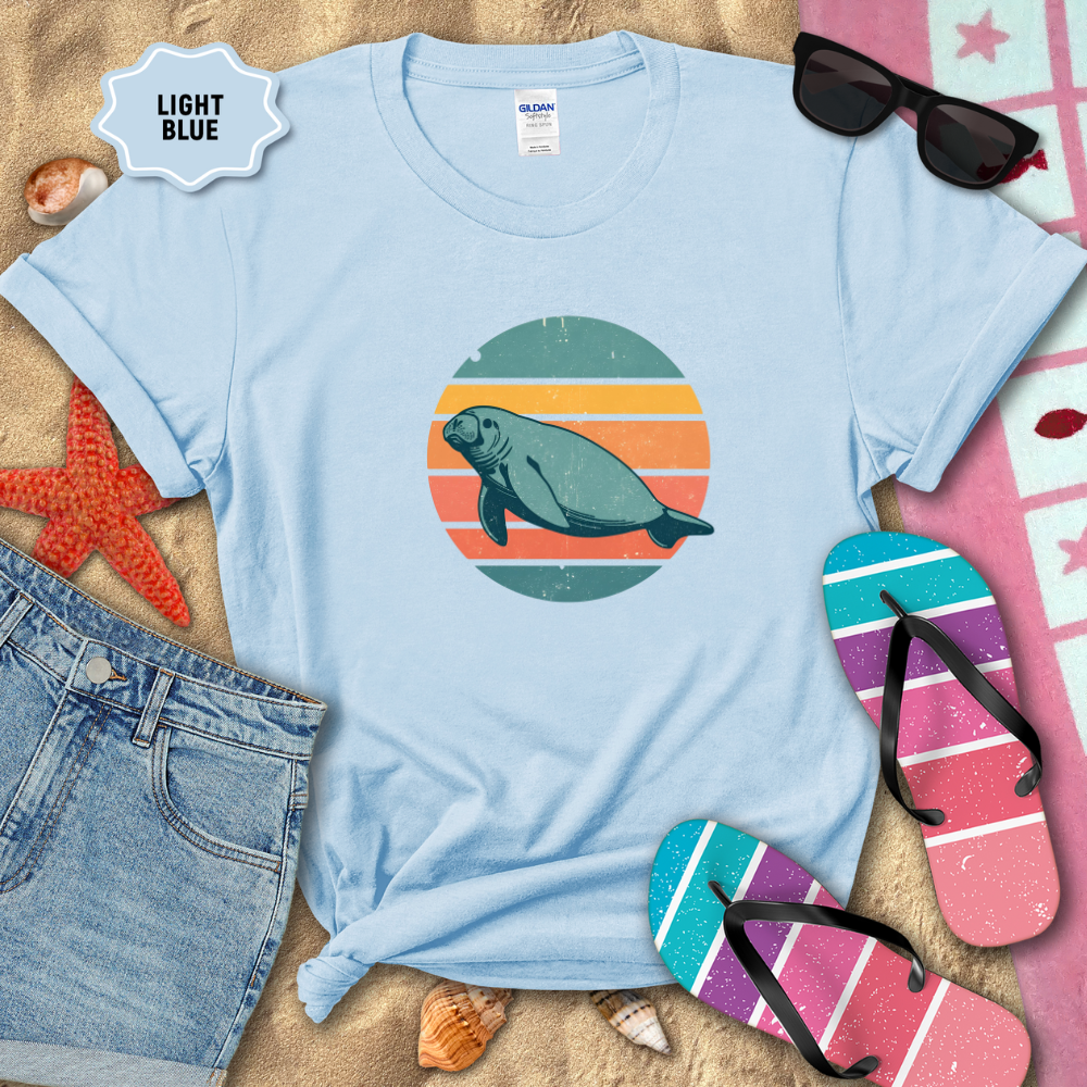 Retro sunset manatee graphic t-shirt in a light blue color, styled on beach sand with seashells, flip-flops, sunglasses, and denim shorts. The shirt features a circular vintage sunset with teal, coral, and orange stripes and a teal manatee illustration in the center.