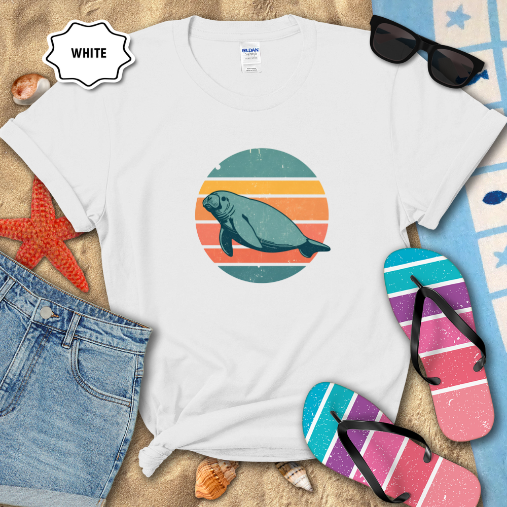 “Retro sunset manatee graphic t-shirt in white, styled on beach sand with seashells, flip-flops, sunglasses, and denim shorts. The shirt features a circular vintage sunset with teal, coral, and orange stripes and a teal manatee illustration in the center.
