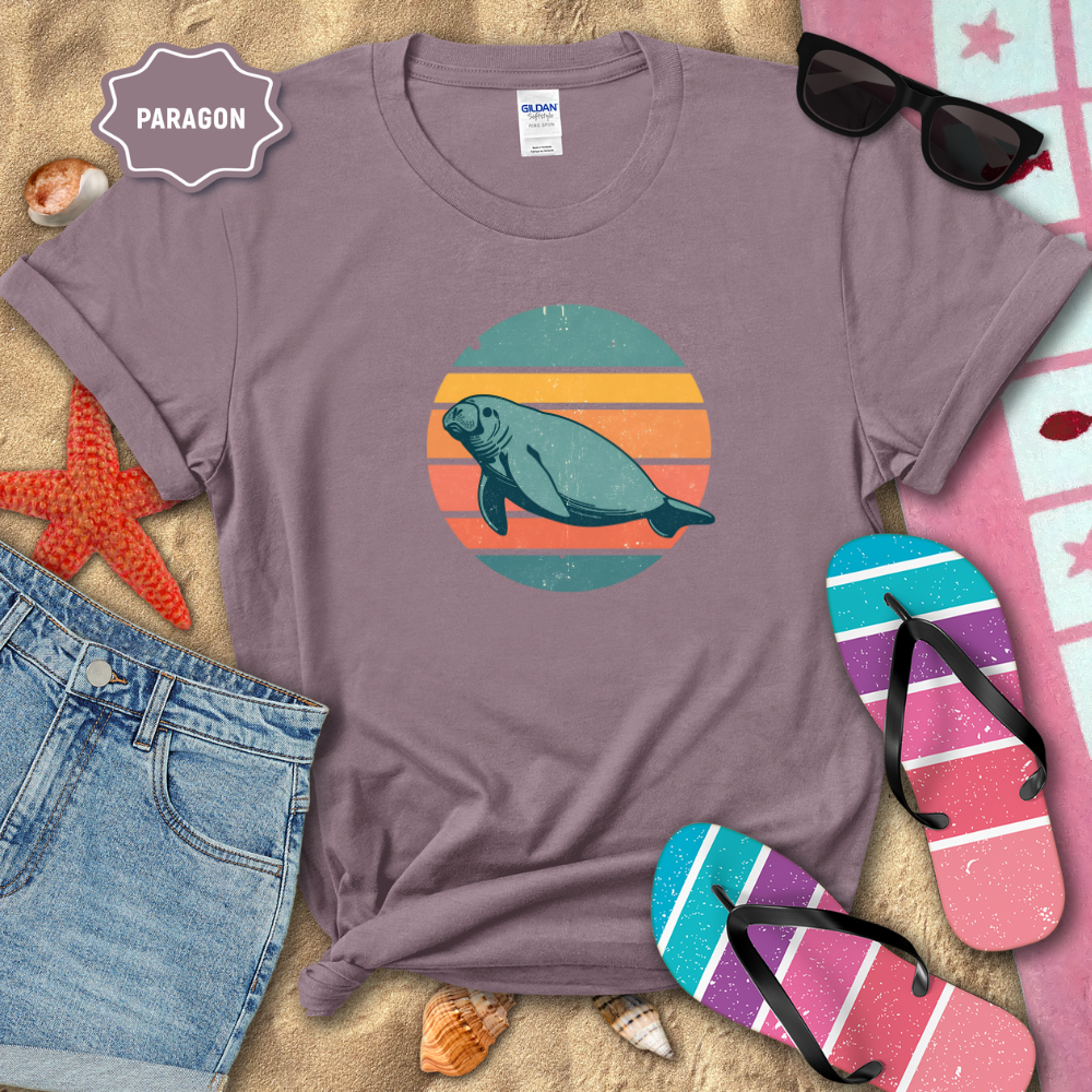 “Retro sunset manatee graphic t-shirt in a muted mauve color, styled on beach sand with seashells, flip-flops, sunglasses, and denim shorts. The shirt features a circular vintage sunset with teal, coral, and orange stripes and a teal manatee illustration in the center.