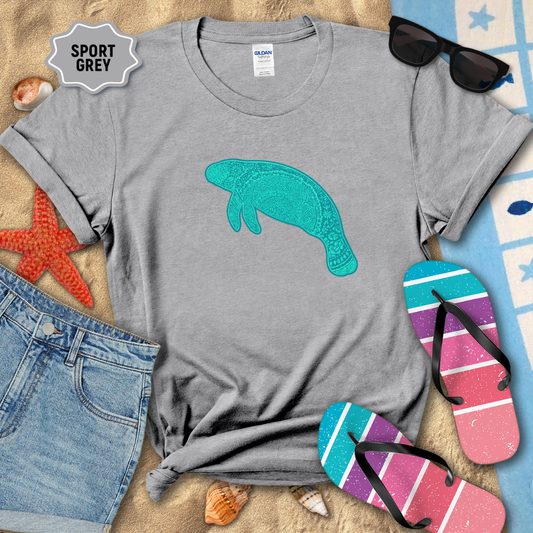 Sport Grey unisex t-shirt laid flat on beach sand with a teal mandala manatee graphic printed on the front. The shirt is styled with denim shorts, colorful striped flip-flops, seashells, a starfish, and sunglasses.