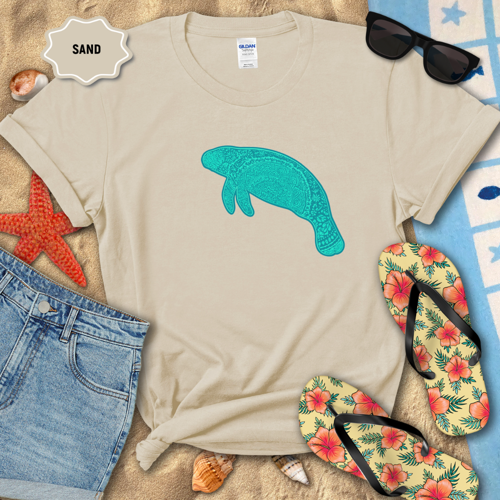 Sand colored unisex t-shirt laid flat on beach sand with a teal mandala manatee graphic printed on the front. The shirt is styled with denim shorts, colorful striped flip-flops, seashells, a starfish, and sunglasses.