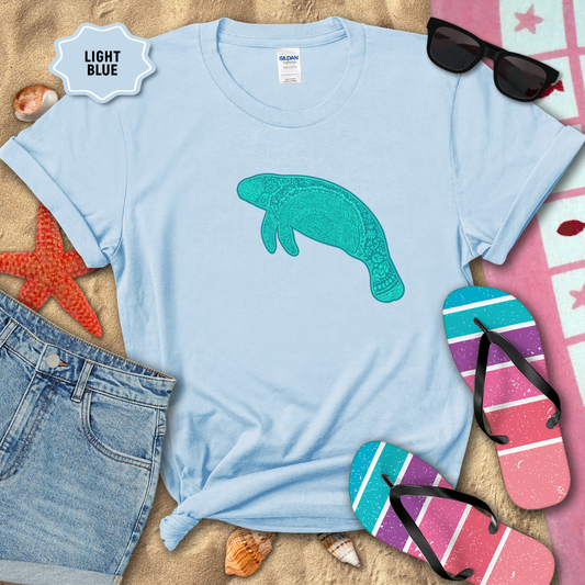 Light Blue unisex t-shirt laid flat on beach sand with a teal mandala manatee graphic printed on the front. The shirt is styled with denim shorts, colorful striped flip-flops, seashells, a starfish, and sunglasses.
