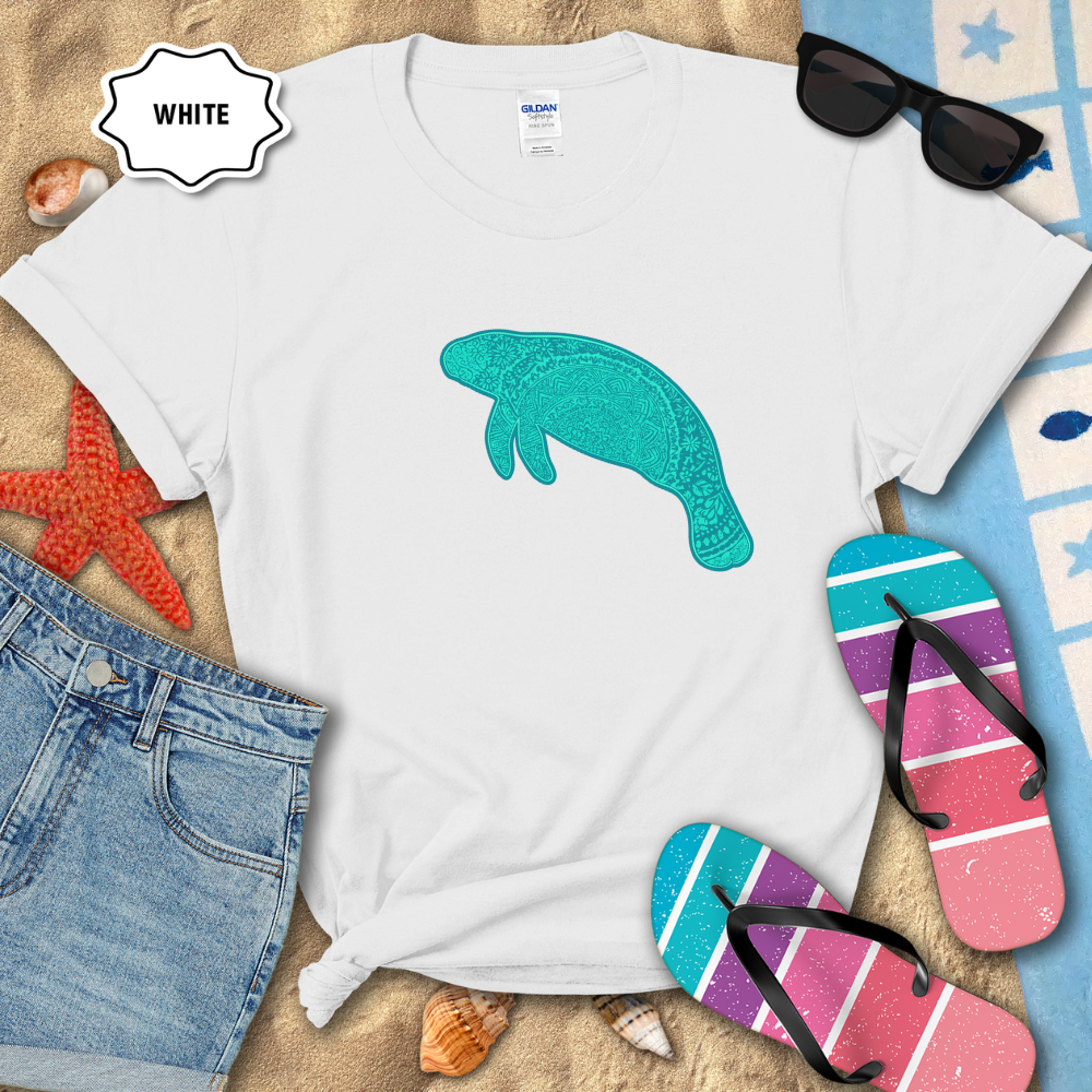 White unisex t-shirt laid flat on beach sand with a teal mandala manatee graphic printed on the front. The shirt is styled with denim shorts, colorful striped flip-flops, seashells, a starfish, and sunglasses.