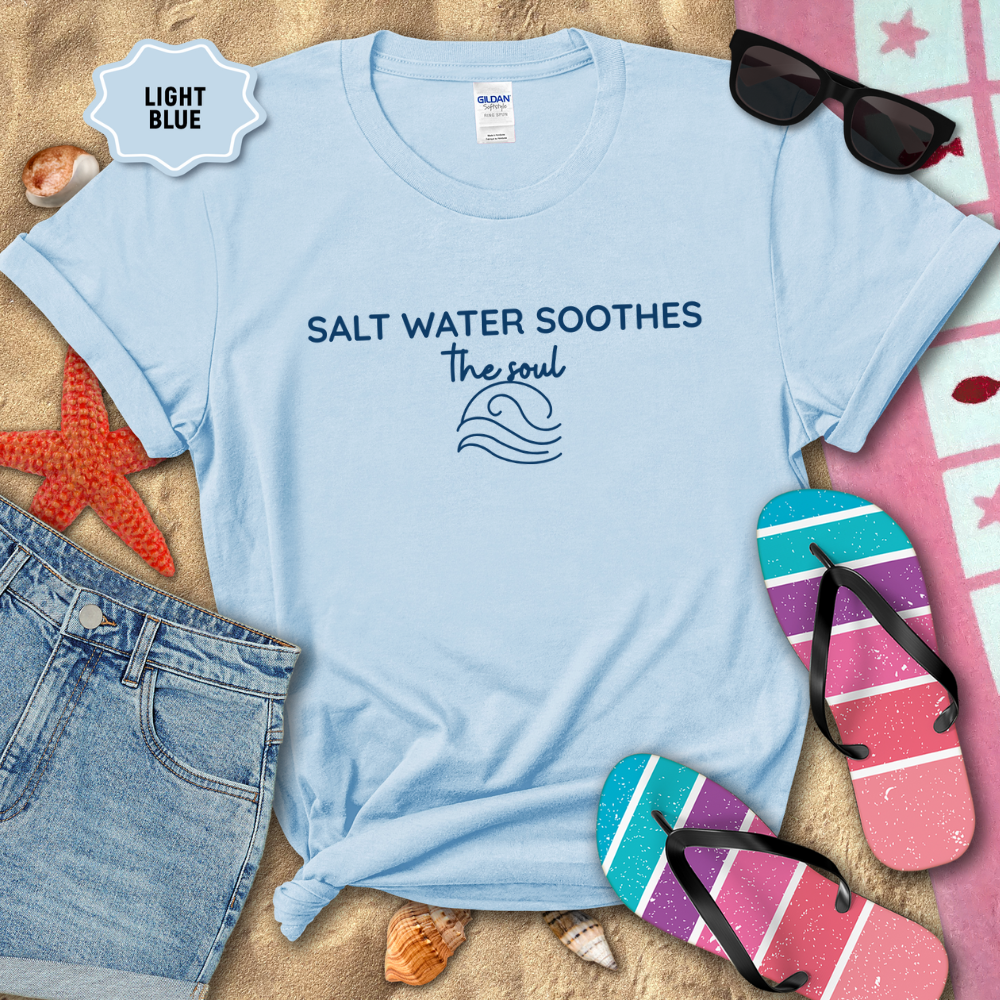 Light blue coastal t-shirt laid flat on sand with the phrase ‘Salt Water Soothes the Soul’ and a minimalist wave graphic in navy. Styled with denim shorts, colorful flip-flops, sunglasses, seashells, and a starfish for a beach-themed aesthetic.