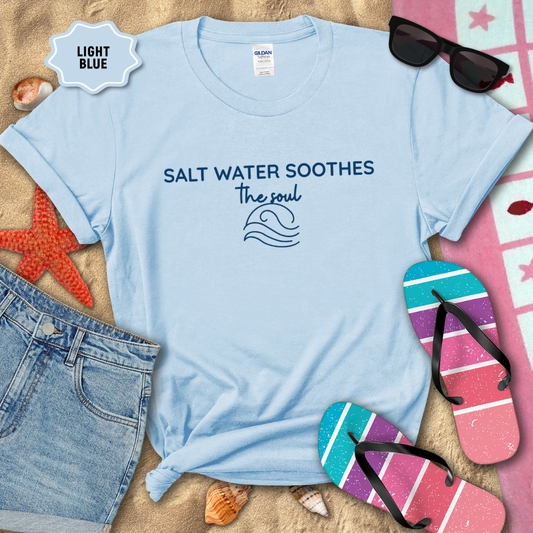 Light blue coastal t-shirt laid flat on sand with the phrase ‘Salt Water Soothes the Soul’ and a minimalist wave graphic in navy. Styled with denim shorts, colorful flip-flops, sunglasses, seashells, and a starfish for a beach-themed aesthetic.