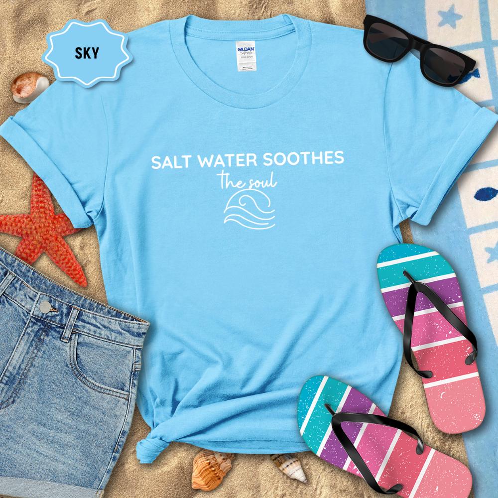 Sky Blue  coastal t-shirt laid flat on sand with the phrase ‘Salt Water Soothes the Soul’ and a minimalist wave graphic in navy. Styled with denim shorts, colorful flip-flops, sunglasses, seashells, and a starfish for a beach-themed aesthetic.