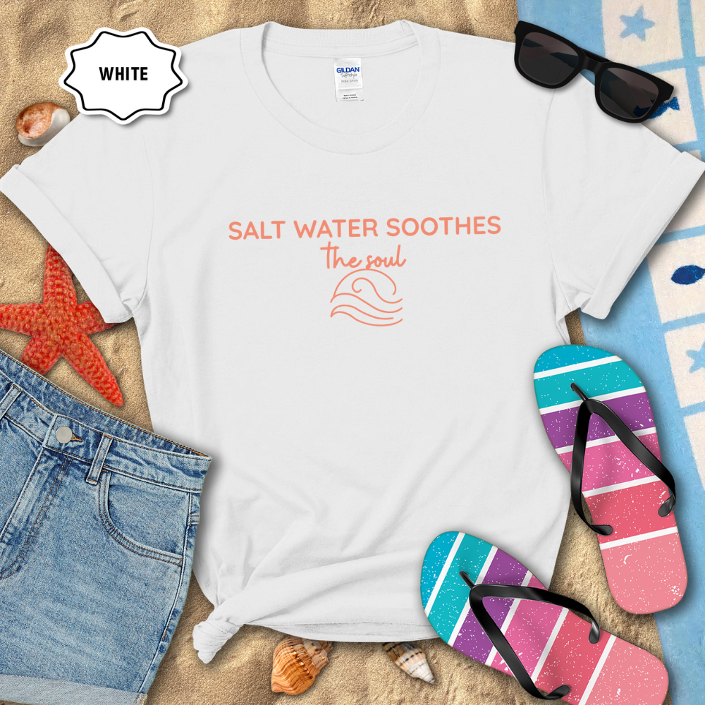 White coastal t-shirt laid flat on sand with the phrase ‘Salt Water Soothes the Soul’ and a minimalist wave graphic in navy. Styled with denim shorts, colorful flip-flops, sunglasses, seashells, and a starfish for a beach-themed aesthetic.