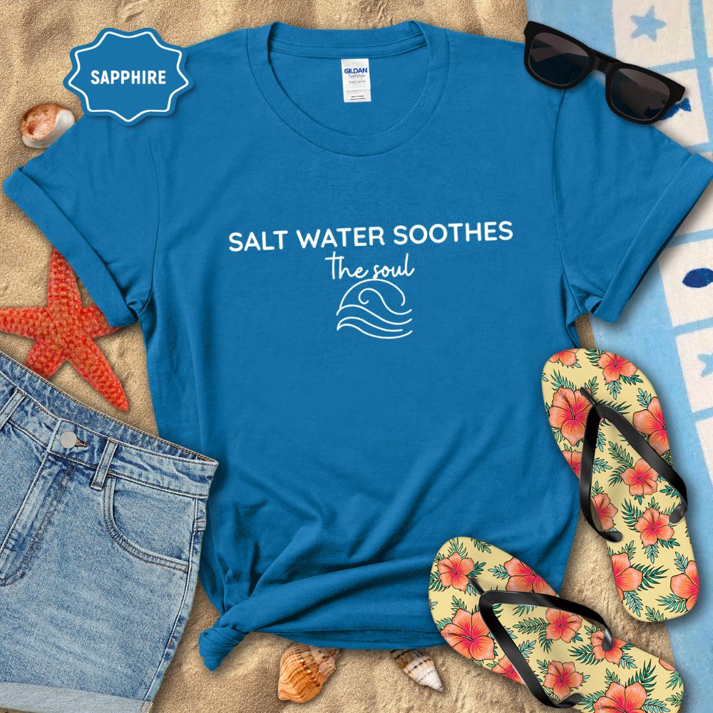 Sapphire colored  coastal t-shirt laid flat on sand with the phrase ‘Salt Water Soothes the Soul’ and a minimalist wave graphic in navy. Styled with denim shorts, colorful flip-flops, sunglasses, seashells, and a starfish for a beach-themed aesthetic.