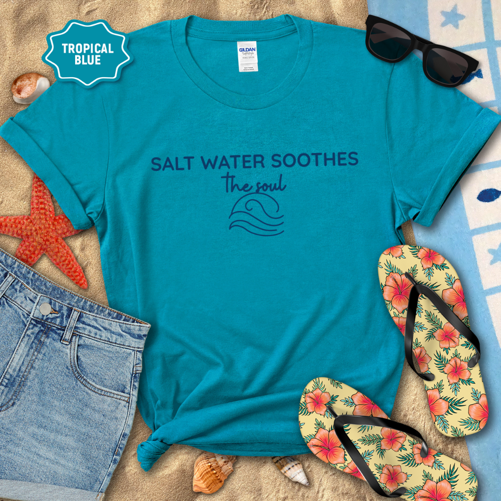 Tropical Blue coastal t-shirt laid flat on sand with the phrase ‘Salt Water Soothes the Soul’ and a minimalist wave graphic in navy. Styled with denim shorts, colorful flip-flops, sunglasses, seashells, and a starfish for a beach-themed aesthetic.