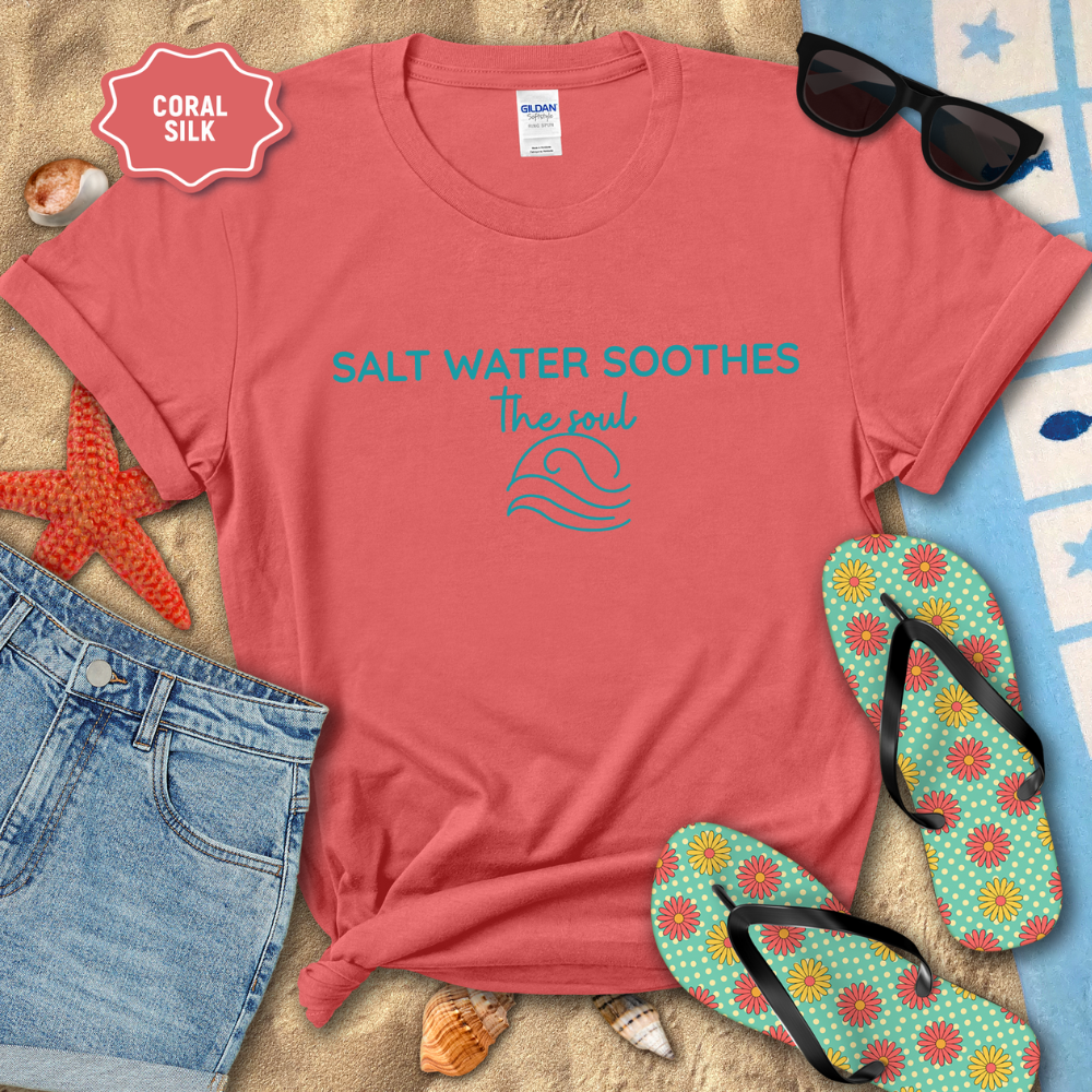 Coral silk coastal t-shirt laid flat on sand with the phrase ‘Salt Water Soothes the Soul’ and a minimalist wave graphic in navy. Styled with denim shorts, colorful flip-flops, sunglasses, seashells, and a starfish for a beach-themed aesthetic.