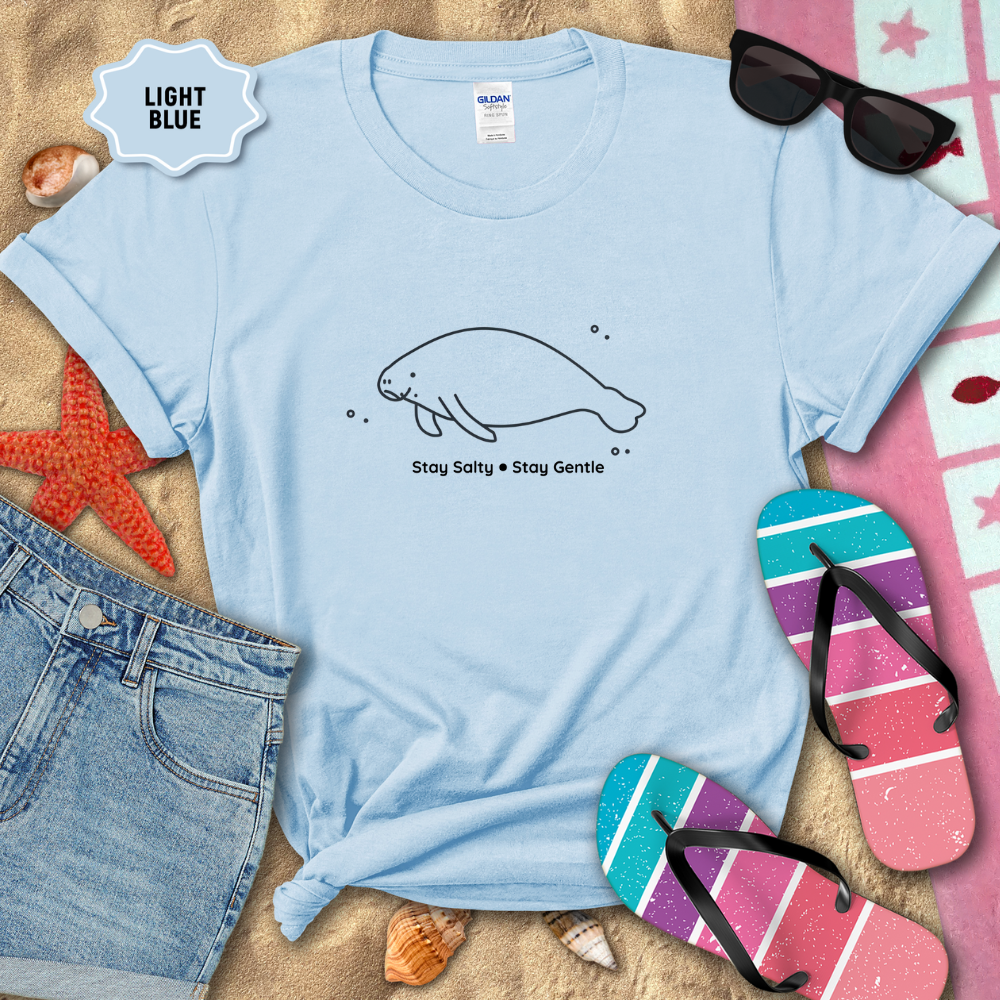 Light blue t-shirt with a manatee design and text, surrounded by beach items on sand.