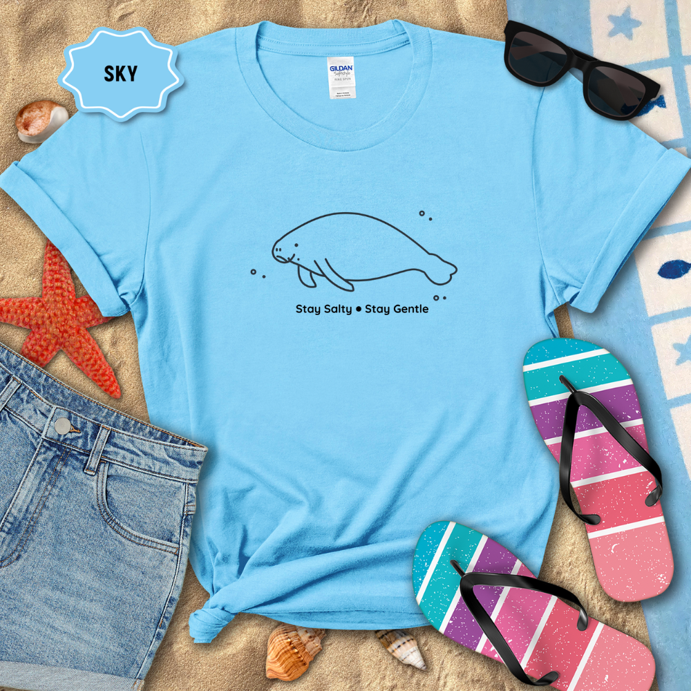 Light blue t-shirt with a manatee design and text, surrounded by beach items on sand.