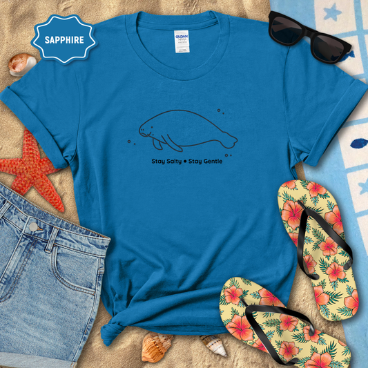 Blue t-shirt with a manatee graphic and text, surrounded by beach accessories on sand.