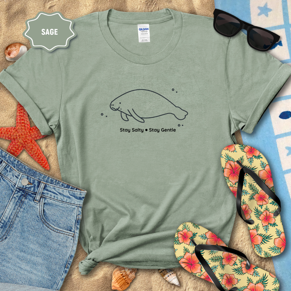 Green t-shirt with a manatee design and text, surrounded by beach accessories on sand.