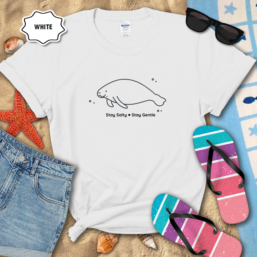 White t-shirt with a manatee design and text, surrounded by beach items on sand.