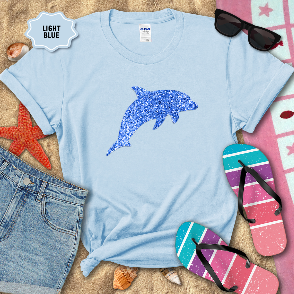 Light blue t-shirt with a dolphin design on a sandy background with beach accessories.