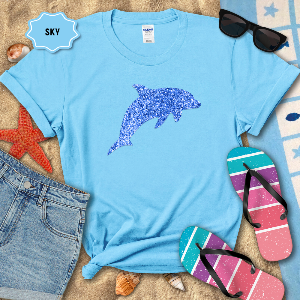 Light blue t-shirt with a dolphin design on a sandy surface with beach accessories.