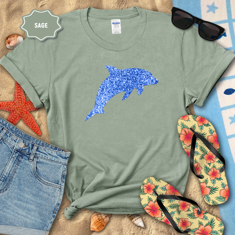Sage green t-shirt with blue dolphin graphic on a sandy surface with beach accessories.
