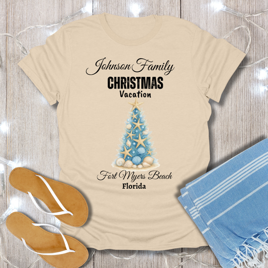 Beige t-shirt with Christmas tree design and text on a wooden surface with flip-flops and a towel.