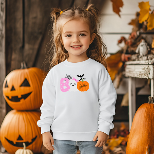 Child wearing a white sweatshirt with Halloween-themed design in front of pumpkins and autumn decorations.