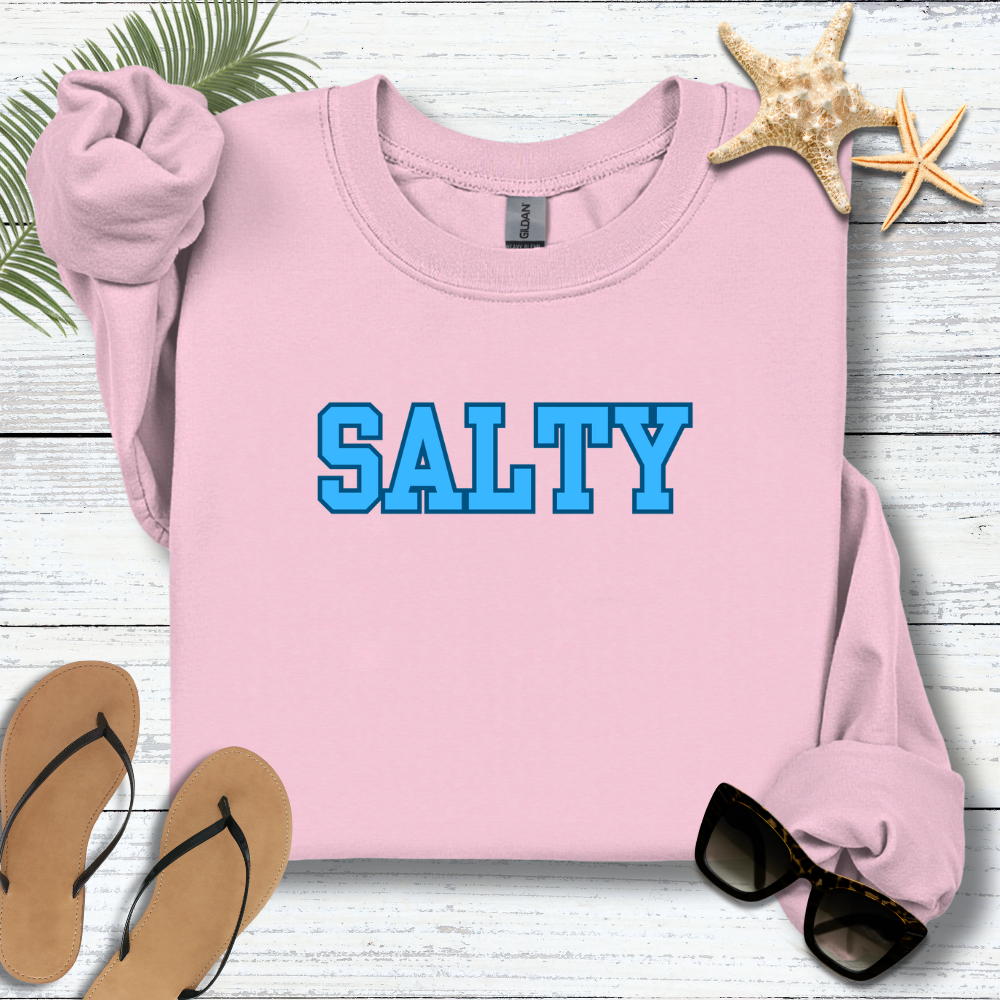 Pink sweatshirt with 'SALTY' in blue text on a wooden surface with flip-flops and sunglasses.