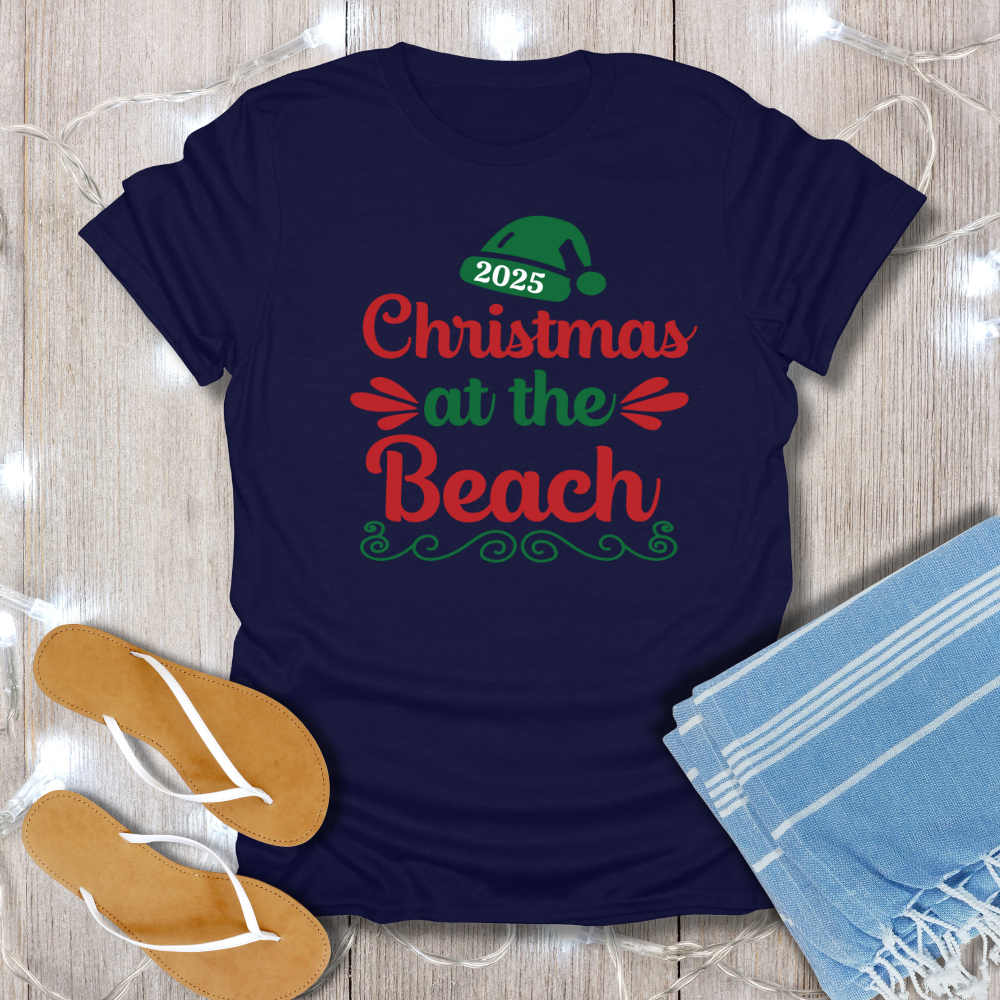 Navy blue t-shirt with '2025 Christmas at the Beach' text on a wooden surface with flip-flops and a towel.