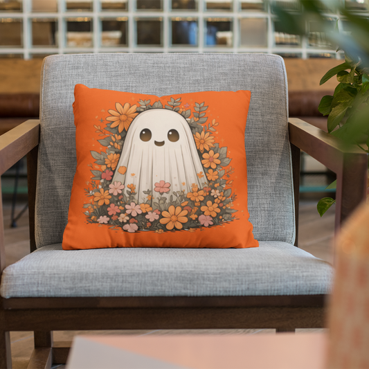 Bright orange throw pillow featuring a smiling cartoon ghost surrounded by soft fall-colored flowers, placed on a modern gray chair in a cozy room.