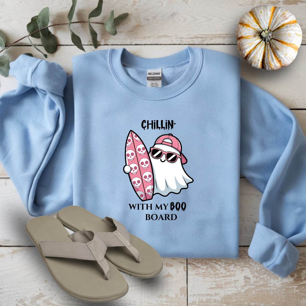 Light blue sweatshirt with ghost and surfboard graphic on a wooden surface with flip-flops and a small pumpkin.