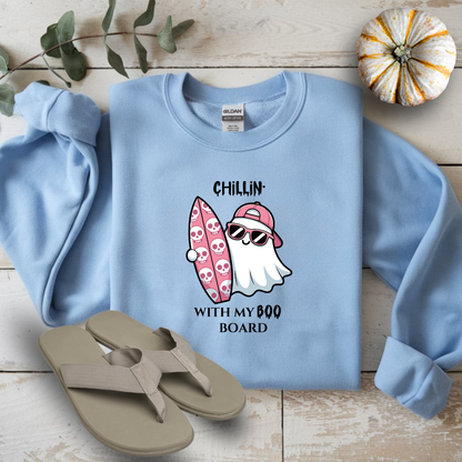 Light blue sweatshirt with ghost and surfboard graphic on a wooden surface with flip-flops and a small pumpkin.