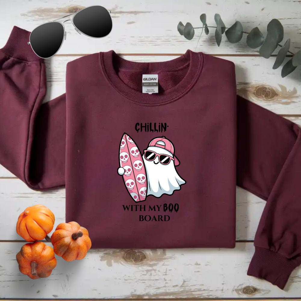 Maroon sweatshirt with ghost and surfboard graphic on a wooden surface with pumpkins and sunglasses.
