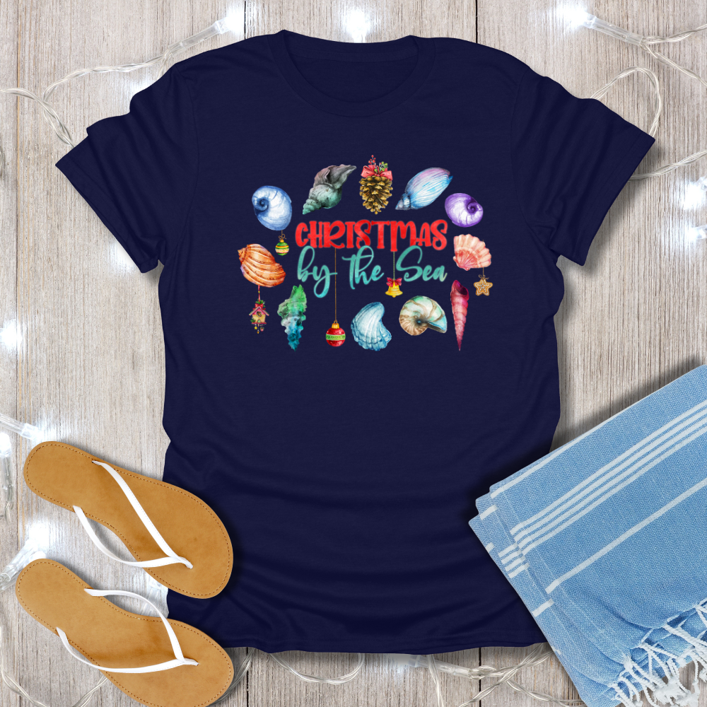 Navy blue t-shirt with 'Christmas by the Sea' design on a wooden surface with flip-flops and a towel.
