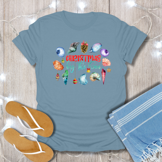 Blue t-shirt with Christmas-themed design on a wooden surface with flip-flops and a towel.