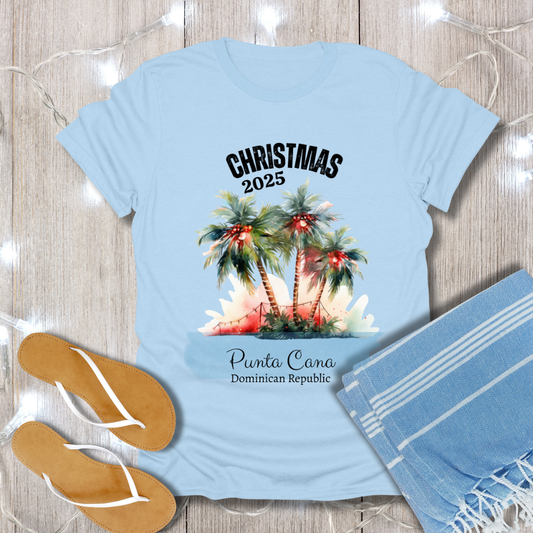 Light blue t-shirt with Christmas design featuring palm trees and 'Christmas 2025 Punta Cana Dominican Republic' text, placed on a wooden surface with flip-flops and a towel.