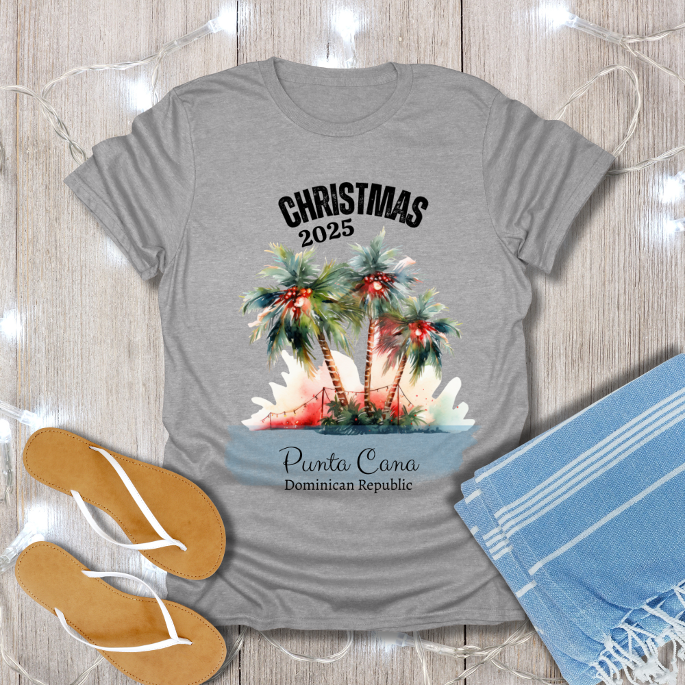 Gray t-shirt with Christmas 2025 Punta Cana design on a wooden surface with flip-flops and a towel.