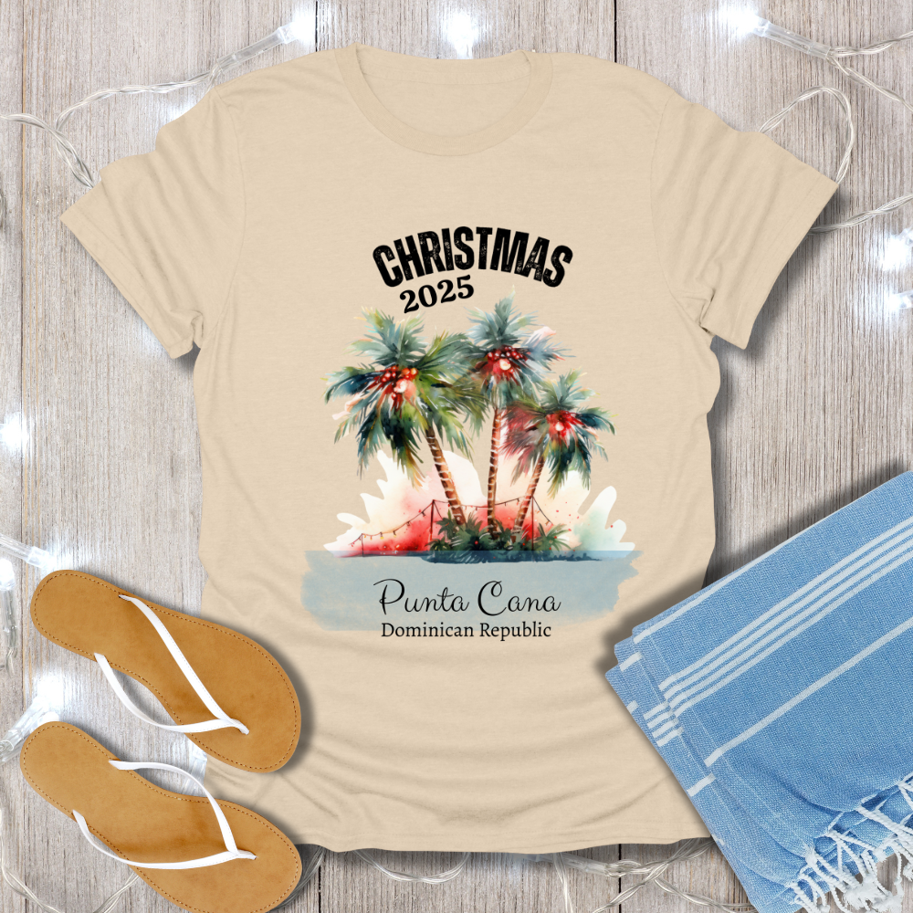 Beige t-shirt with Christmas 2025 Punta Cana Dominican Republic design on a wooden surface with flip-flops and a towel.