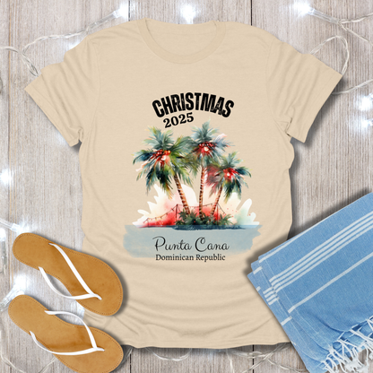 Beige t-shirt with Christmas 2025 Punta Cana Dominican Republic design on a wooden surface with flip-flops and a towel.