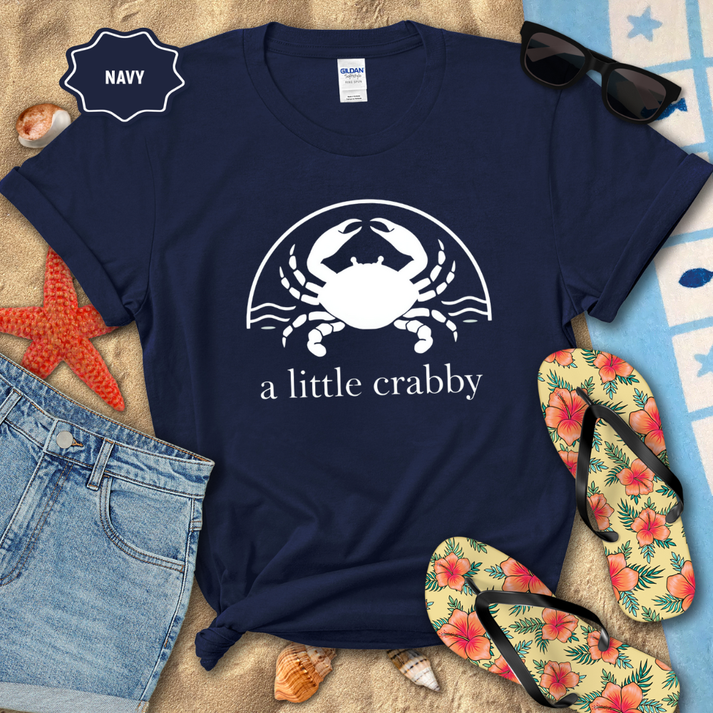 Navy t-shirt with a crab graphic and text, surrounded by beach accessories on sand.