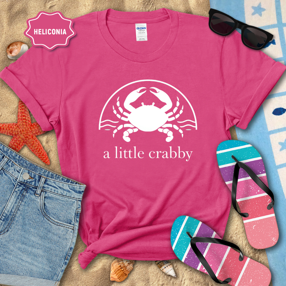 Pink t-shirt with a crab design and text, sunglasses, and flip-flops on a beach-themed background.