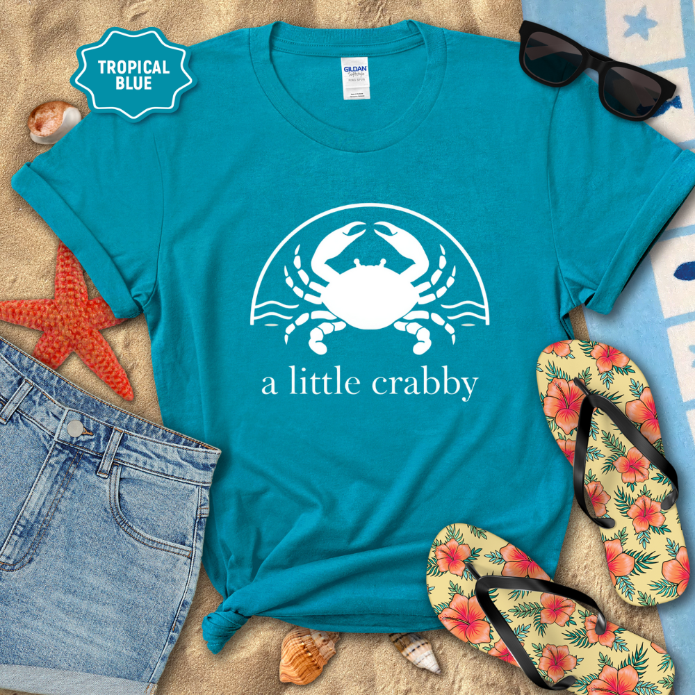 Blue t-shirt with a crab graphic and 'a little crabby' text, surrounded by beach accessories on sand.