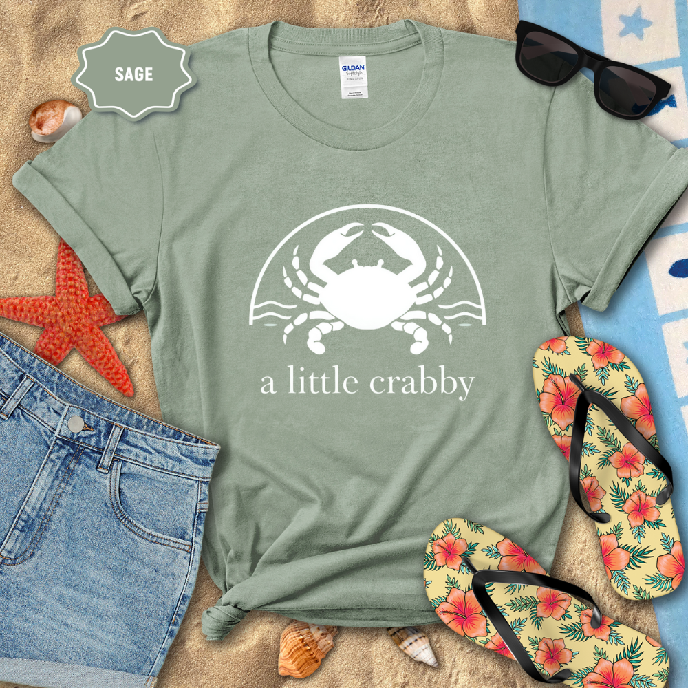 Sage green t-shirt with a crab graphic and 'a little crabby' text, surrounded by beach accessories.