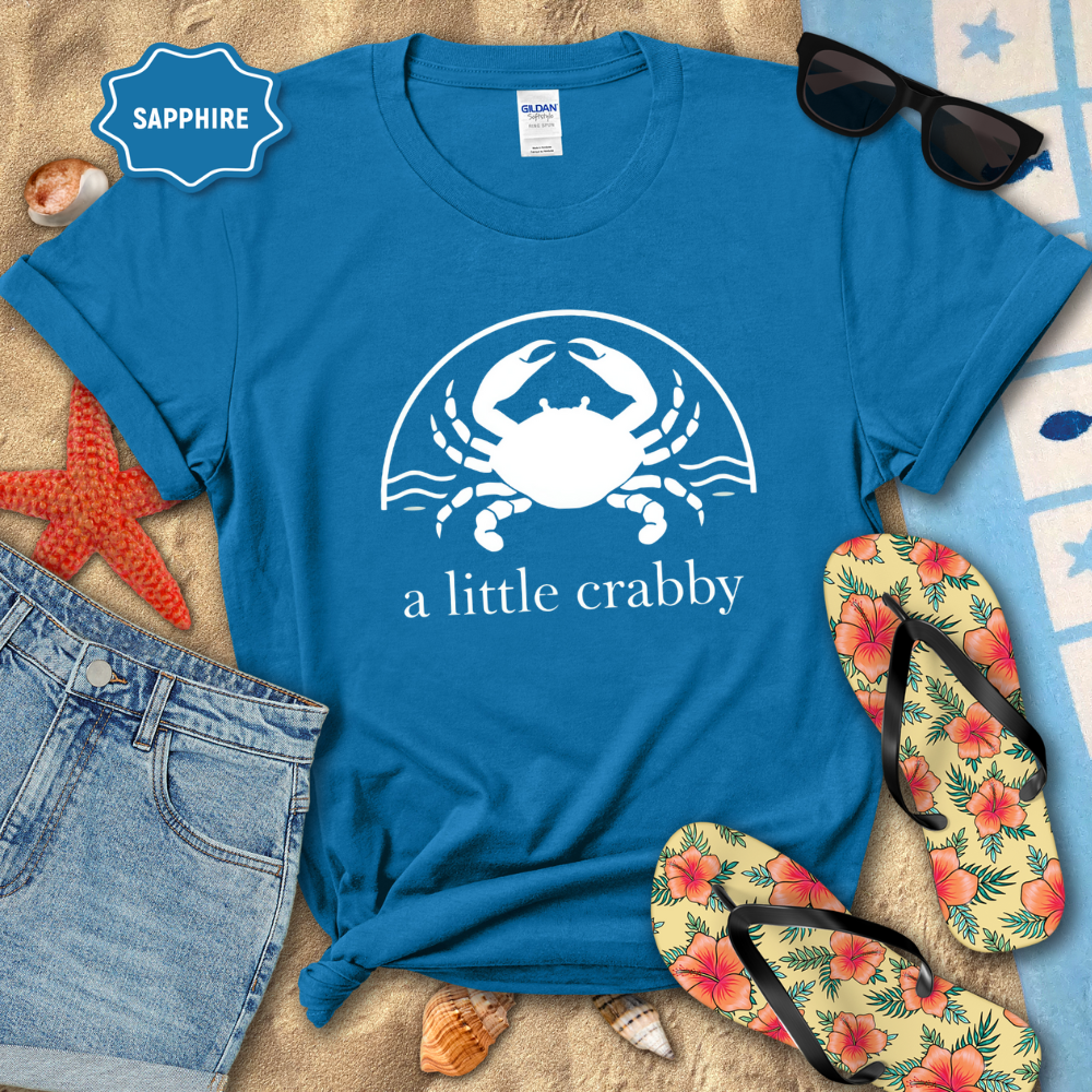 Blue t-shirt with a crab graphic and text, surrounded by beach accessories on sand.
